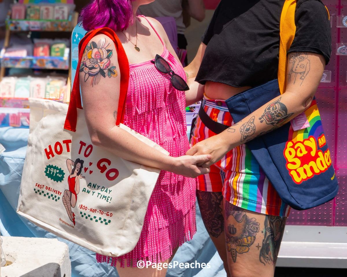 Two women with colorful tattoos and accessories are standing together, one holding a white bag with a red and green design and the other carrying a blue bag with a rainbow pattern.