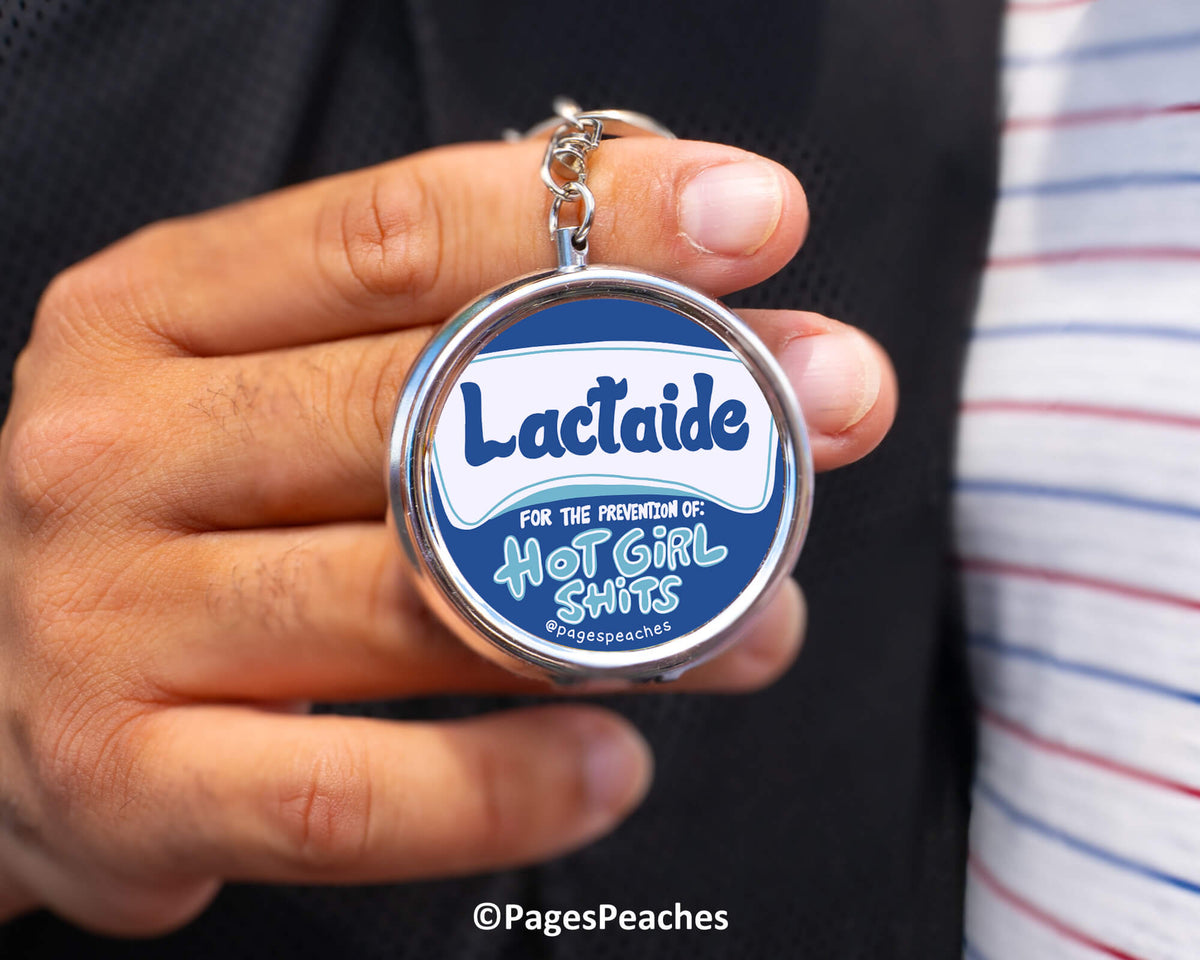 A person is holding a keychain with a circular design featuring the text "Lactaid" and "For the prevention of hot girl shitss" along with a graphic of a hot girl.
