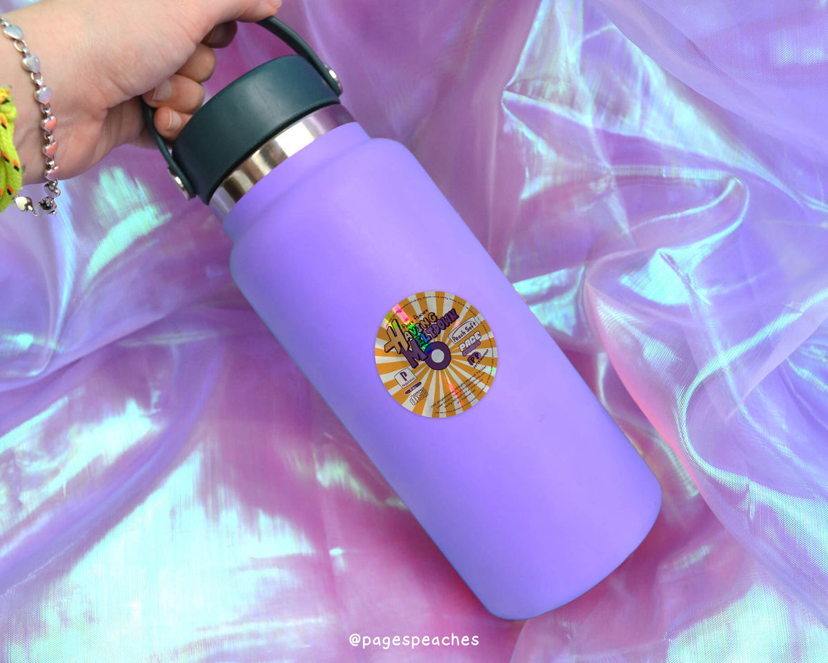 A purple water bottle with a sticker on it, resting on a pink fabric background.