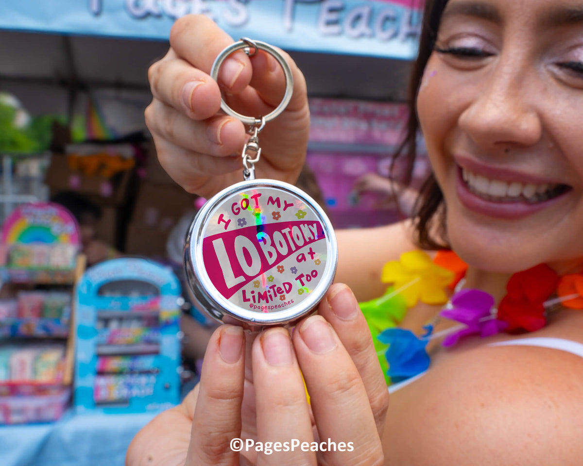 A woman is holding a round keychain with the text "I got my LOBOTY limited edition" on it, smiling and wearing colorful lei garland.