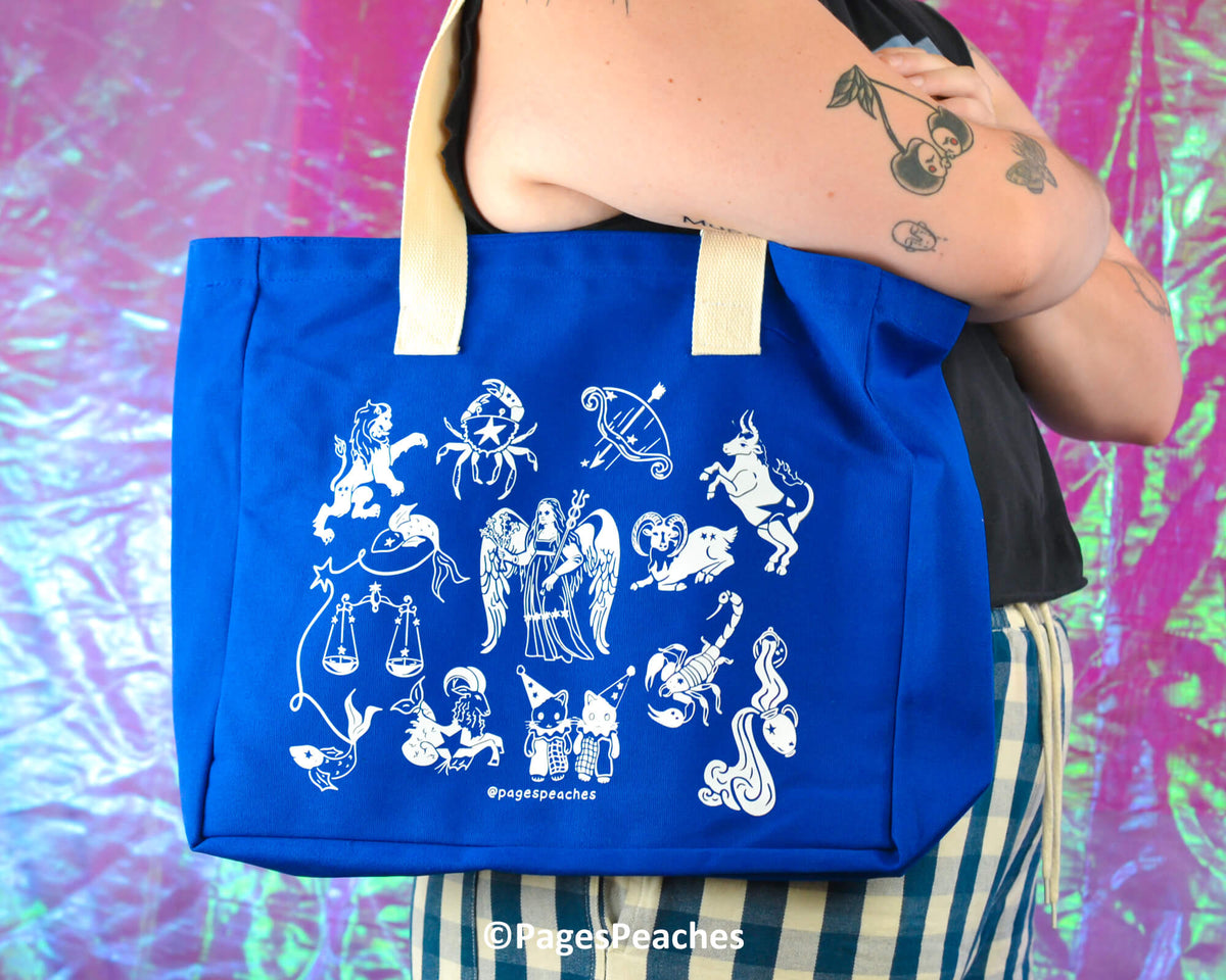 Blue tote bag with white zodiac designs held by a person against a colorful background