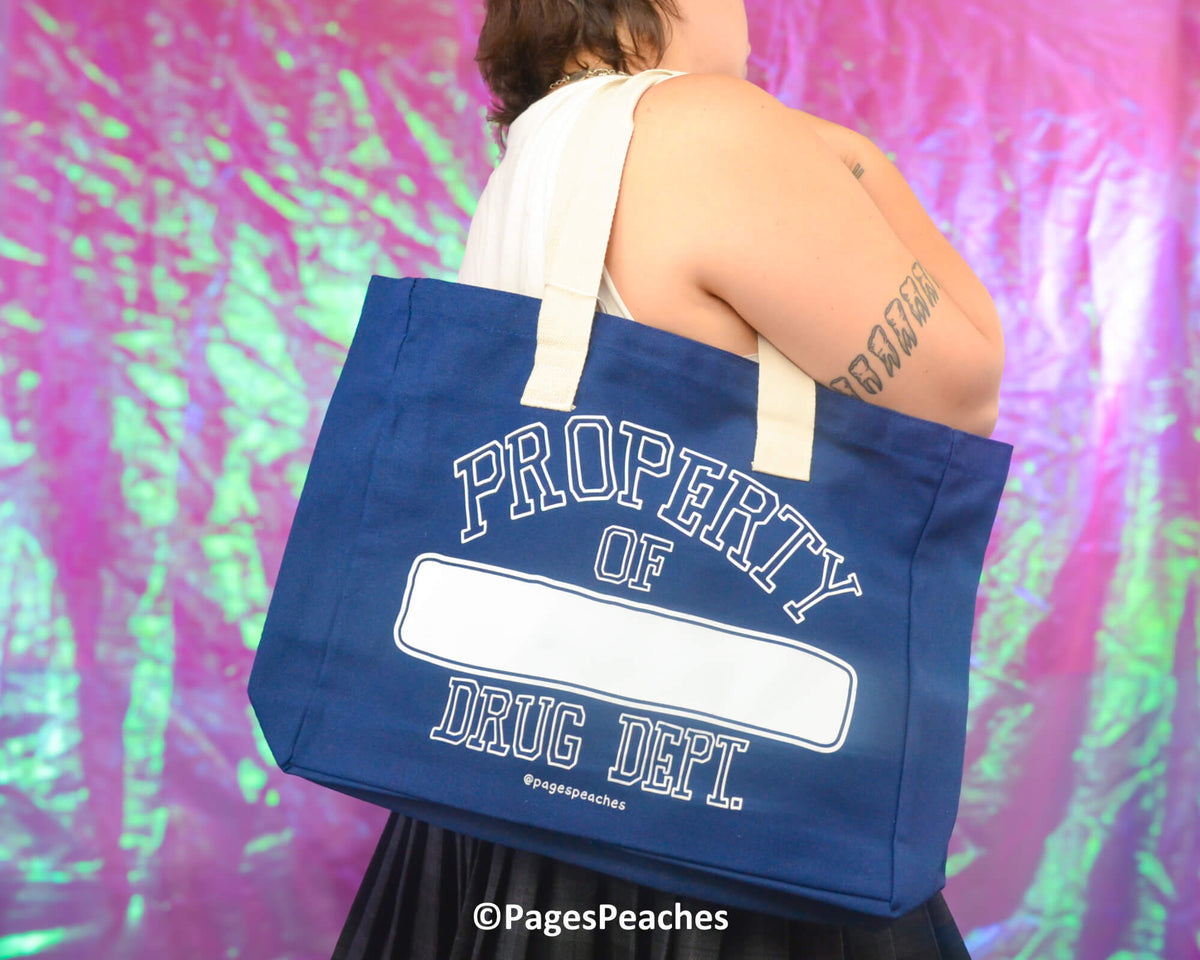 Person holding a blue tote bag with 'Property of Drug Dept.' text against a colorful, abstract background.