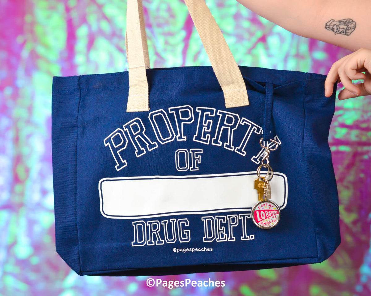 Blue tote bag with 'Property of Drug Dept' text and keychain, held by a person against a colorful background.
