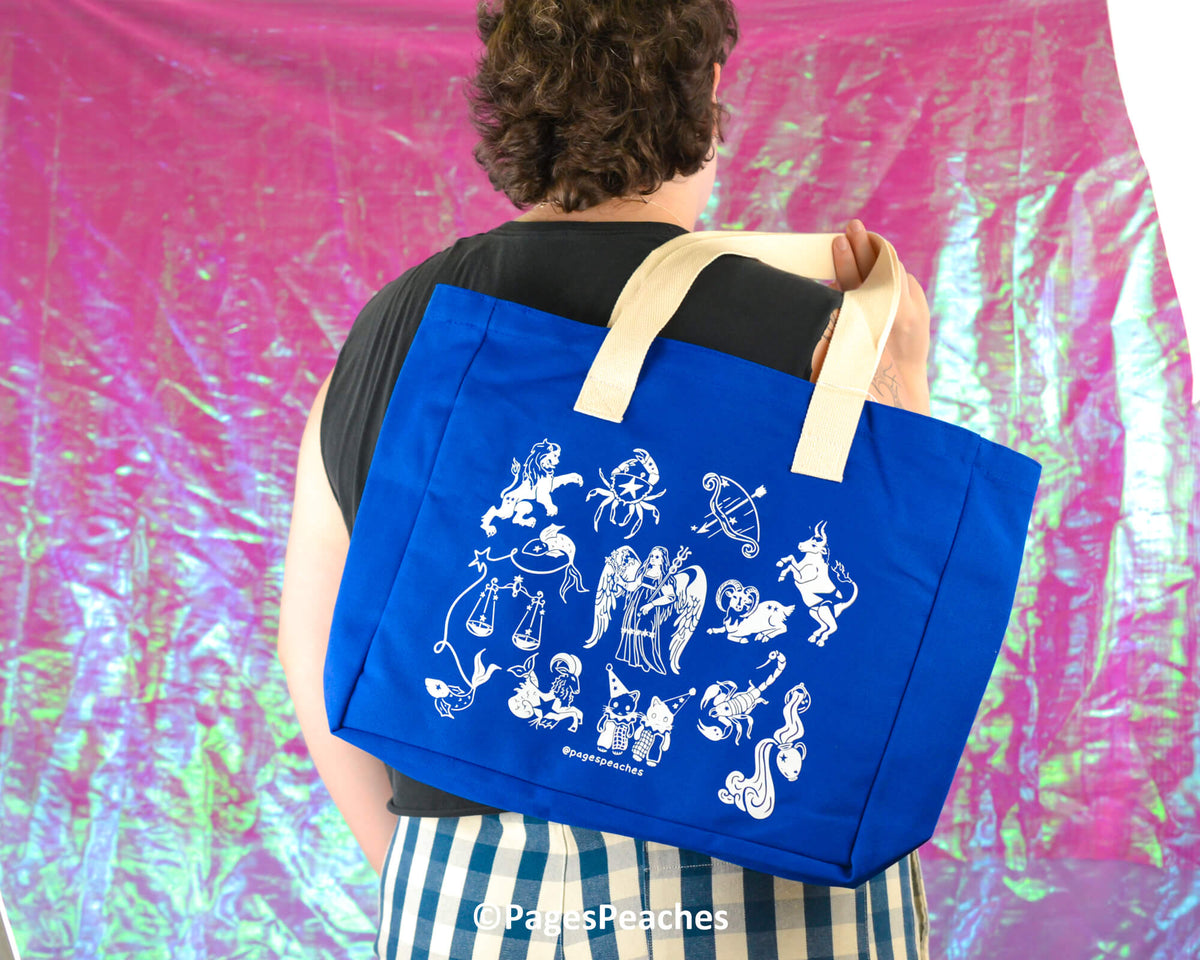 Person holding a blue tote bag with white zodiacs against a colorful background