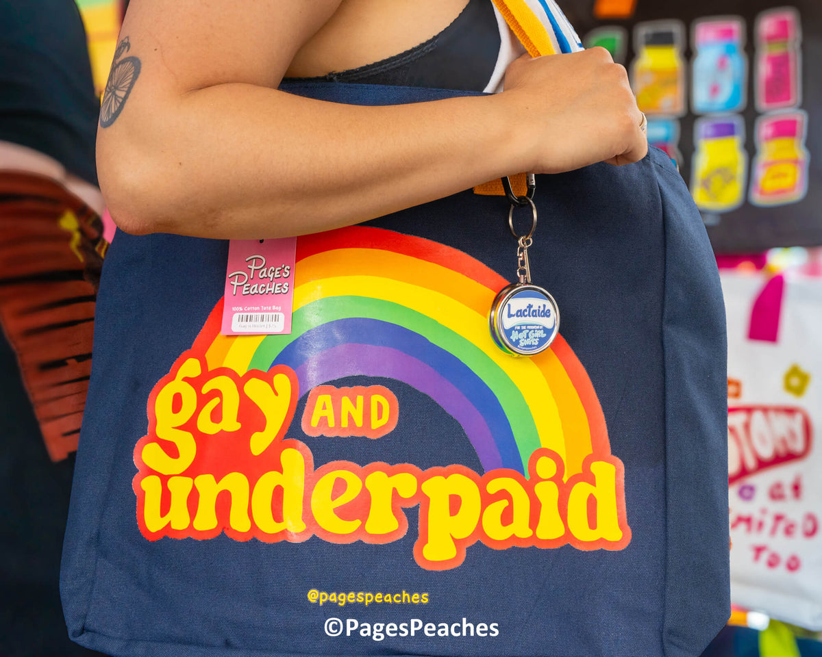 A person is holding a blue tote bag with a rainbow graphic and the text "Gay and underpaid" printed on it.