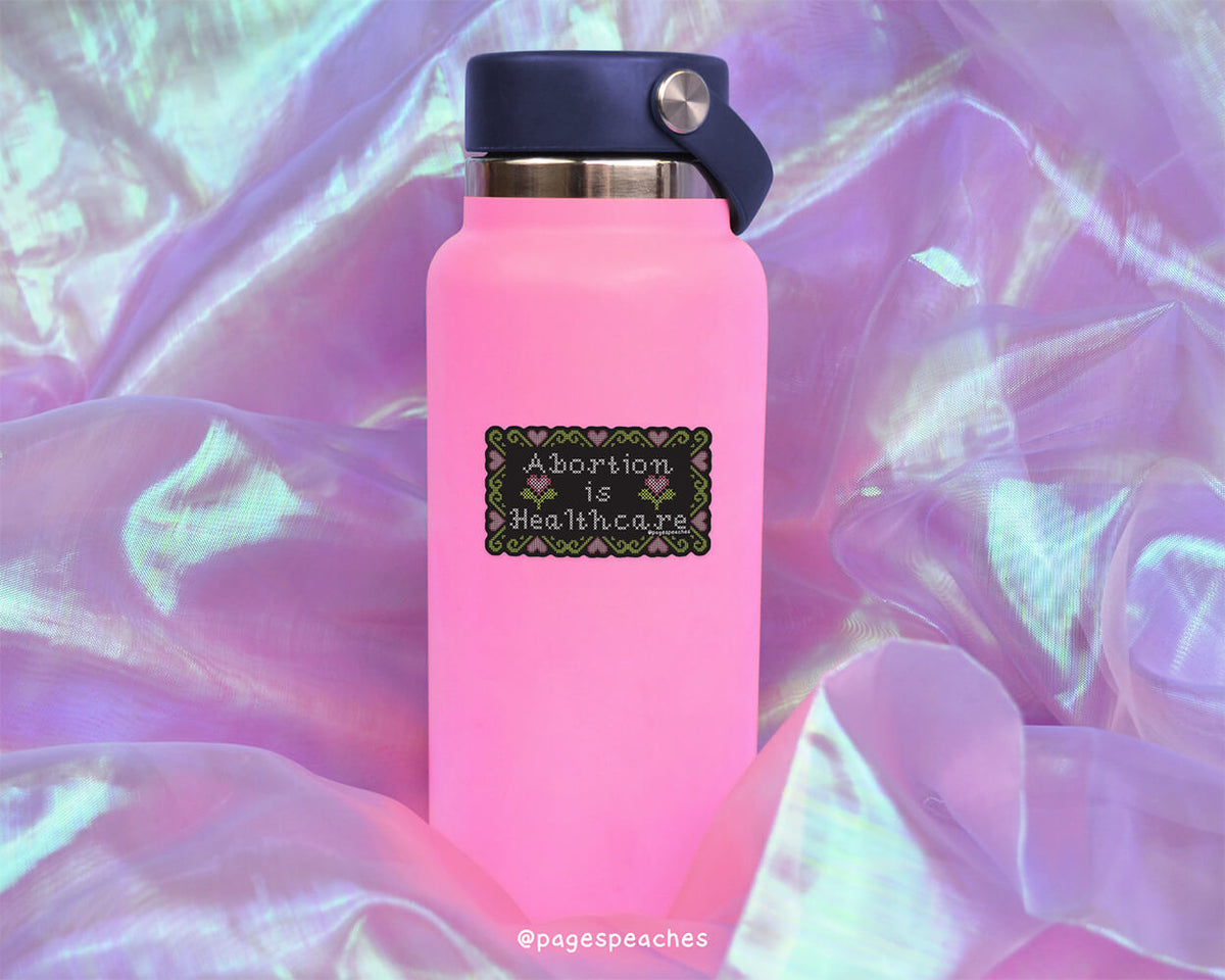 A pink water bottle with a black and white sticker that says "Abortion is healthcare" against a purple and green background.