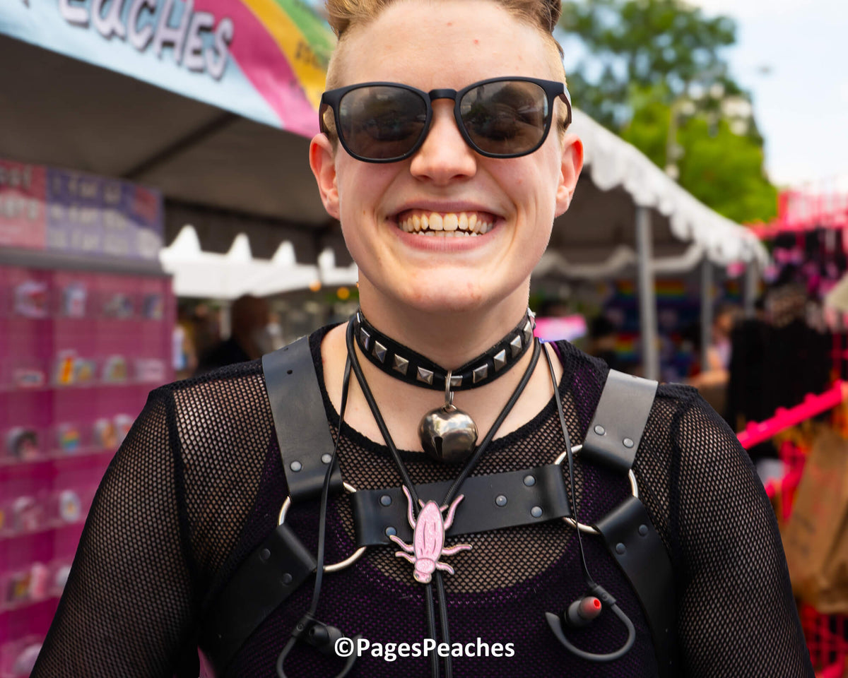 A smiling person wearing sunglasses, a studded choker, and a bell necklace, standing in front of a pink and white tent.