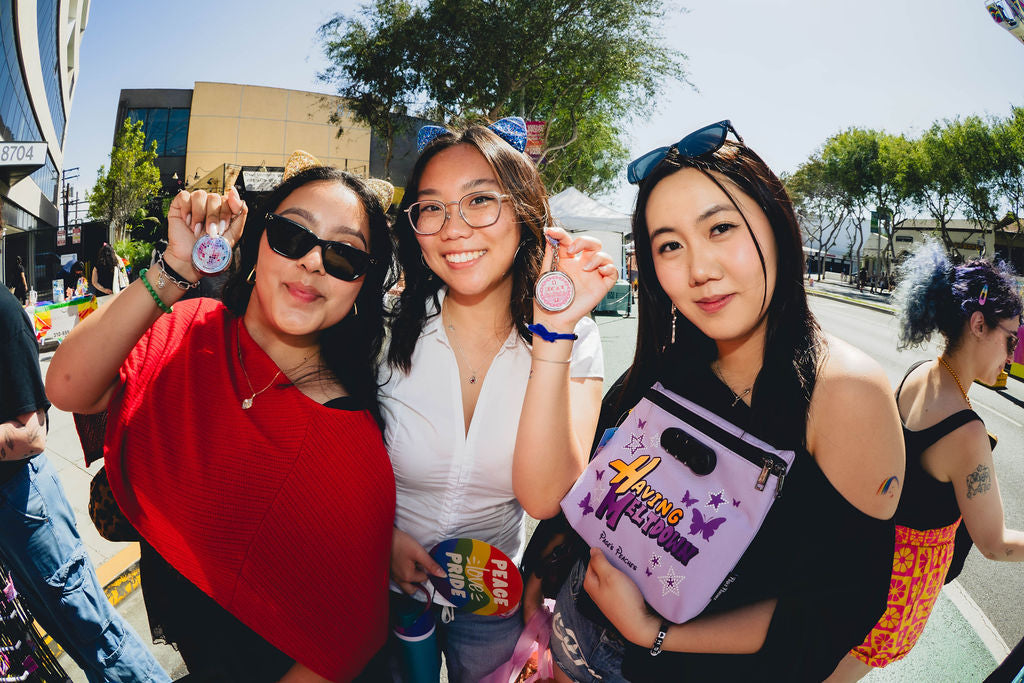 Three young women posing together outdoors, each holding a small object, such as a badge or a sign, and smiling at the camera.