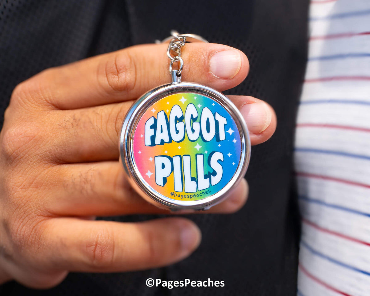 A person is holding a keychain with the text "FAGGOT PILLS" and a rainbow-colored design.