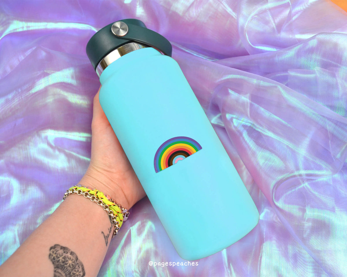 A person's hand holding a blue water bottle with a rainbow sticker on it, against a purple fabric background.