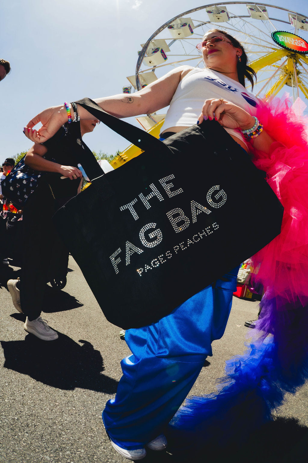 A woman wearing a white top and holding a large black bag with the text "THE FAG BAG" printed on it, standing in front of a ferris wheel.