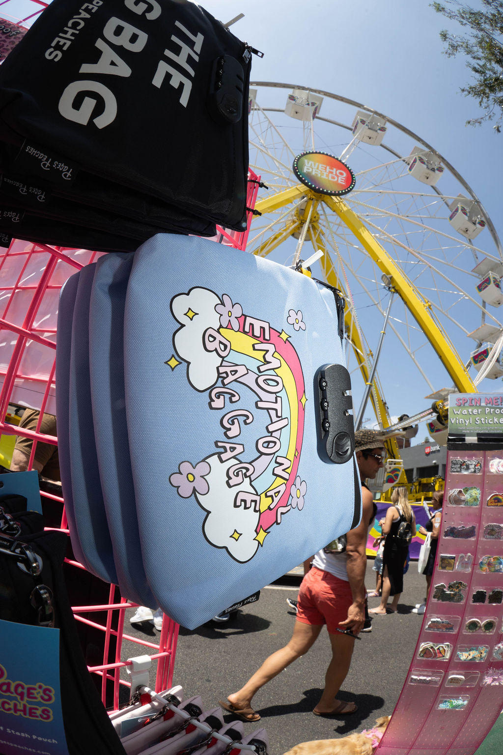 a colorful and vibrant scene at a fairground, with a Ferris wheel and various items for sale, including a large blue bag with a rainbow design and a black bag with the text "THE BIG BAG".