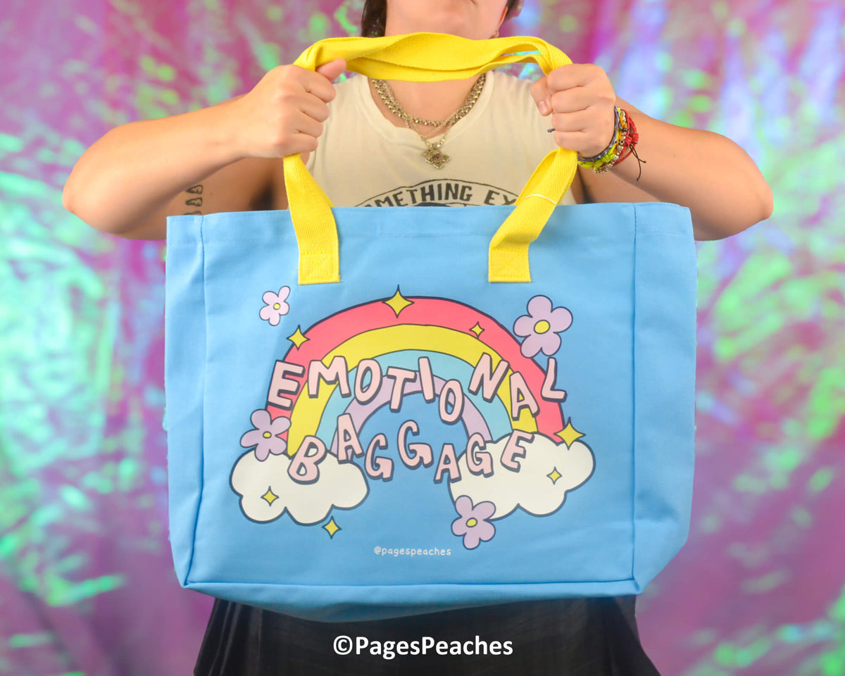 Person holding a blue tote bag with a rainbow design and 'Emotional Baggage' text against a colorful background.