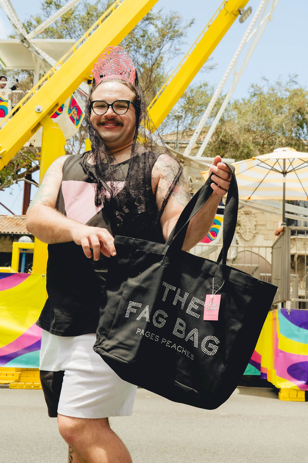 A man wearing a pink crown and glasses is holding a large black bag with the text "THE FAG BAG" printed on it.